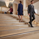 A man in a dark gray suit and a woman in a navy blue coat ascend a grand hotel staircase, with a hotel bellhop in a maroon uniform standing at the top.