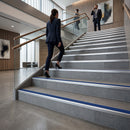 A woman in a dark navy suit walks up a modern gray tiled staircase with glass railings and wood handrails inside a corporate building. Two men in suits are visible further up the stairs in a real indoor environment.