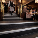 A set of modern black stone stairs with brushed silver metal handrails in a luxurious restaurant or hotel lobby, with a waiter walking up the stairs and patrons seated in the background.