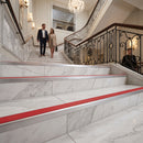 A grand marble staircase in an elegant building, featuring a bright red stripe along the steps, with a man and woman walking up the stairs and a uniformed attendant seated near the railing.