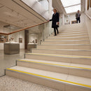 A woman in a dark coat walks up a wide indoor staircase in a modern gallery or museum, with another person further ahead. The stairs have light beige steps with bright yellow striping and metal handrails.