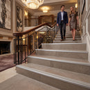 A man and a woman walk down a grand indoor staircase in a luxurious hotel or mansion lobby. The staircase features light gray stone steps with silver metal trim, a dark wood handrail with black metal balustrades, and elegant wall art and chandeliers in the background.