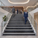 A woman in a navy blue business suit walks down a modern marble staircase in a bright, contemporary office lobby with glass railings and other professionals in the background.