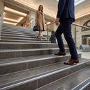 A man in a navy suit and brown dress shoes walks down a grand marble staircase, while a woman in a beige trench coat and black heels carries a handbag up the steps in a luxurious indoor lobby.