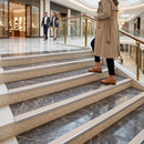 A woman in a beige trench coat and brown loafers walks down a modern marble staircase in a luxury shopping mall, with two men walking in the background.