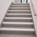 A modern interior staircase with light gray textured steps, white risers, and a brushed metal handrail against off-white walls.