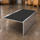 A rectangular metal riser or support bracket with a black top surface and brushed silver aluminum sides, photographed on a concrete warehouse floor.