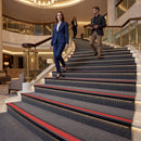 A woman in a navy blue suit walks down a grand hotel staircase with a red stripe, while a man in a dark blazer and khaki pants follows her up the stairs in a luxurious lobby.