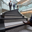 A low-angle view of a carpeted staircase in a modern glass-walled office lobby, with three business people walking up the stairs in the background and a person's brown dress shoe resting on the step in the foreground.