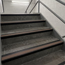 Concrete stair steps with dark brown rubber anti-slip strips and black metal handrails in an indoor stairwell.
