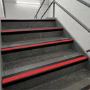 Red and black stair tread strips installed on concrete stairs in a stairwell with white brick walls and metal railings.