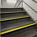 Concrete stairs with bright yellow anti-slip strips and black edging, set in a stairwell with light gray brick walls and metal railings.