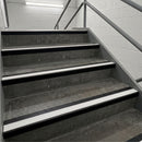 A set of industrial concrete stairs with black rubber anti-slip tread strips and white edge guides, flanked by gray metal handrails against a white brick wall.