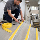 A man kneels on concrete stairs installing bright yellow safety striping alongside metal rails, with a roll of matching yellow strips and tools nearby.