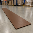 A long, thin rectangular brown board or strip placed diagonally on a gray concrete warehouse floor, with shelving and packaged pallets blurred in the background.