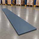 A long, thin rectangular blue-gray board or panel placed diagonally on a polished concrete warehouse floor, with pallets and shelving visible in the background.