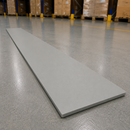 A long, thin rectangular gray board or tile lies diagonally on a polished concrete warehouse floor, with pallets and shelving visible in the blurred background.