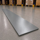 A long, rectangular gray metal sheet or panel lies diagonally on a polished concrete warehouse floor, with pallets and shelving visible in the blurred background.