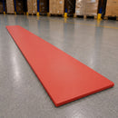 A long, flat red rectangular mat or board placed diagonally on a gray warehouse floor, with pallets and storage racks blurred in the background.