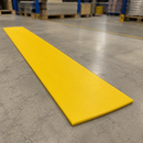 A long, bright yellow rectangular mat or pad placed diagonally on a gray concrete warehouse floor, with blurred industrial shelving and pallets in the background.