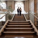A man and a woman walk down a grand indoor staircase with wood steps, glass railings, and warm architectural lighting. The setting appears to be a luxury residential interior.