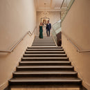 A modern indoor staircase with dark stone steps and warm under-step lighting, flanked by cream walls and metal handrails. Two people are walking down the stairs in the distance, with a chandelier and framed wall art above.