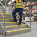 Yellow anti-slip GRP stair nosing fitted to the edges of a concrete warehouse staircase, with a worker in hi-vis safety gear walking down the steps