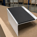 A silver aluminum extrusion profile with a black textured adhesive pad on its top surface, shown on a workbench with a blurred warehouse background.