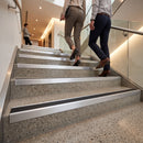 Two people walking up a modern indoor staircase with stainless steel handrails and textured gray stone steps. The staircase features integrated metal grab handles and a black anti-slip strip on each step.
