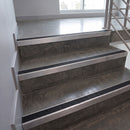 Concrete stairs with brushed metal handrails and black textured non-slip strips, shown in an indoor stairwell with a window and metal railing.