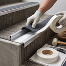 A person wearing white work gloves applies a dark gray ribbed stair carpeting strip to a concrete stair tread, with rolls of matching carpeting tape and installation tools nearby.