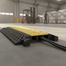 Yellow and black modular cable protector ramp laid across a warehouse floor with power cables routed through its channels and a forklift working in the background