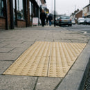 Buff blister tactile paving bonded onto grey pavement at a dropped kerb on a UK high street