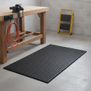 Plain black anti-fatigue rubber mat laid on a workshop floor in front of a wooden workbench with a vice
