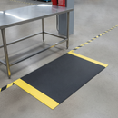 Black anti-fatigue mat with yellow hi-vis safety border in front of a stainless steel factory workstation