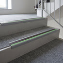 Aluminium bullnose stair nosing fitted to carpet-tiled concrete steps in an office stair core, green photoluminescent strip ahead of a dark anti-slip insert