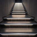 Aluminium LED stair nosing with black anti-slip insert fitted to a dark tiled cinema staircase, riser diffusers lit warm white