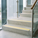 Twin-insert rakeback aluminium stair nosing with high-visibility yellow anti-slip inserts fitted along concrete steps in a glazed stairwell