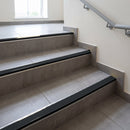 Wide 52mm aluminium tile-in stair nosing with Black RAL 9011 ribbed insert on a grey tiled stairwell, anchoring leg bedded under the tile