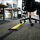 Low profile two channel rubber cable cover with yellow strip running across office carpet tiles as a chair castor rolls over it