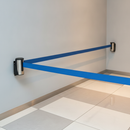 Stainless steel wall-mounted retractable belt post with a blue belt extended across a tiled floor area at a retail queue entrance