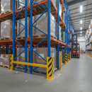 Yellow heavy-duty steel pallet racking upright protector bolted to the floor at the base of a loaded warehouse racking aisle, guarding against forklift impacts