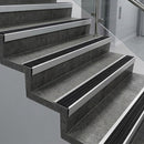Raked back nosings with black castellated inserts on dark concrete steps beside a glass balustrade