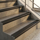Black anodised stair nosings edging sandstone-effect steps in a sunlit stairwell with a steel handrail