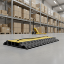 Black and yellow 3-channel cable protector ramp laid across a warehouse aisle floor with a power cable running through it, a yellow pallet truck and racking with boxes in the background