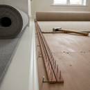 Carpet gripper rods nailed along a skirting board during a carpet installation, with underlay and rolled carpet nearby