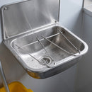 Close-up inside the bowl of a wall mounted cleaners sink showing the removable stainless wire bucket grating and the 38mm flush grated waste outlet, with water droplets across the brushed steel