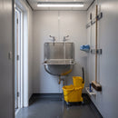 Stainless steel cleaners bucket sink wall mounted in a school janitorial cupboard, a chrome lever bib tap running water into the bowl above a yellow mop bucket on a wringer trolley, with a flat mop and broom on the wall rail alongside