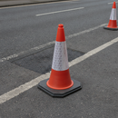 Orange PVC traffic cone with reflective sleeve standing on a tarmac road next to a repaired pothole patch, with a second cone marking the roadworks area