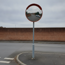 Convex traffic mirror with a red hood on a galvanised pole at a UK car park exit, reflecting the access road and a parked car